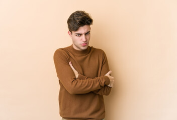 Young caucasian man isolated on beige background frowning face in displeasure, keeps arms folded.