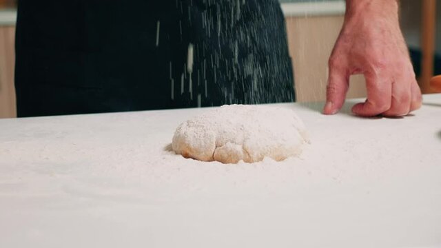 Hands of cook sprinkling flour on pastry dough on the table. Elderly senior baker with bonete and kitchen uniform sieving, sifting, spreading rew ingredients by hand, baking homemade pizza and bread.