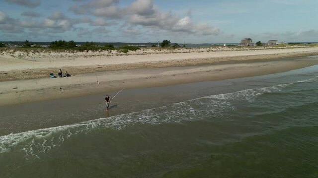 Aerial action shot of a man reeling in a fish with a 14ft fishing rod in slow motion with a bird flying through the scene on an idyllic beach with waves crashing on a semi-cloudy day