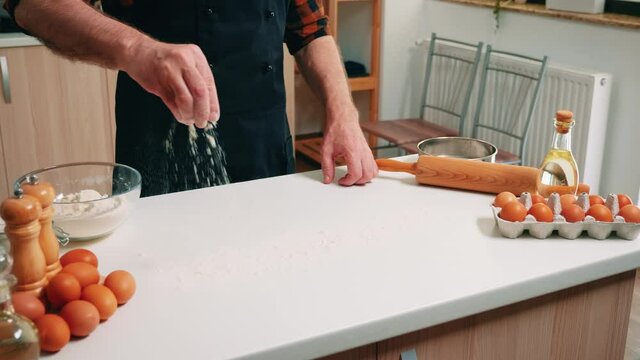 Making dough by man hands at home in kitchen using wheat flour. Retired senior baker with bonete and apron sprinkling, sifting, spreading rew ingredients baking homemade pizza and bread.