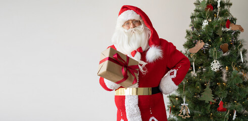 Santa Claus stands near the Christmas tree, holds a box with a gift in his hands and gives it.