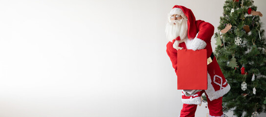 Santa Claus stands near the Christmas tree holding a large paper bag and takes a gift from it.