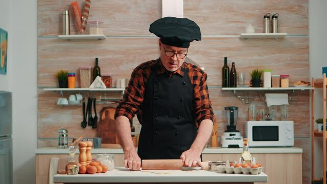 Mature man preparing homemade pizza at home kitchen. Happy elderly chef with bonete using wooden rolling pin kneading raw ingredients to baking traditional cookies, sprinkling, sieving flour on table.