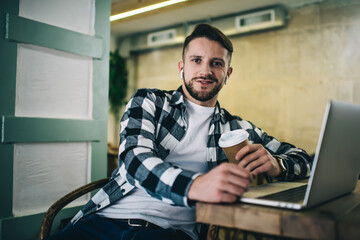 Fototapeta premium Smiling male hipster with takeaway coffee and laptop in cafe