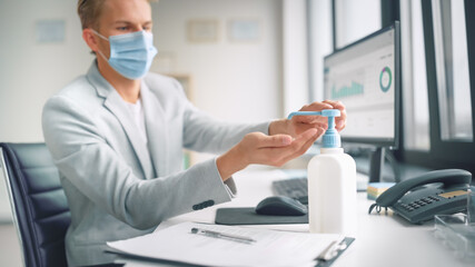 Handsome Stylish Young Man Wearing Protective Face Mask Uses Hand Sanitizer while Working at His Desk on a Computer in Office. Blue Eyed Blond Haired Young Male Follows Health and Safety Instructions