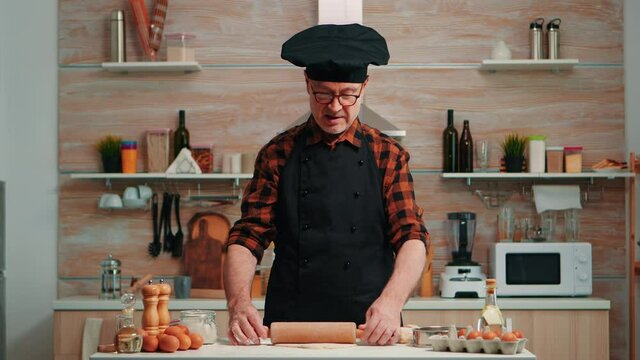 Elderly man rolling dough at home infront of video camera explaining recipe step by step. Retired blogger chef influencer using internet technology communicating on social media with digital equipment