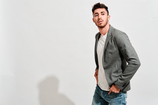 Young man wearing jeans, a white tshit iand a grey blazer walking  in front of a white wall whit his hands inside the jeans pockets