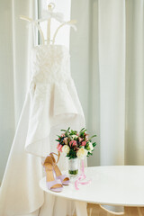 Lilac sandals on a white table with a bouquet of flowers by the window with curtains and a wedding dress on a hanger with a bow tie in the room.