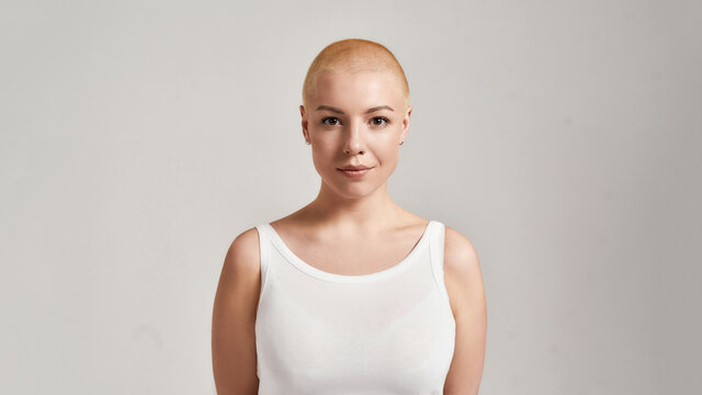 Portrait of a beautiful young caucasian woman with shaved head wearing white shirt, looking at camera while posing isolated over grey background