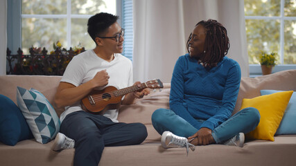 Young romantic multiethnic couple playing ukulele on couch in living room
