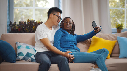 Obraz premium Young diverse joyful couple hugging and smiling while taking selfie photo in living room