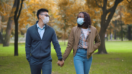 Multiethnic couple in protective masks holding hands walking in park