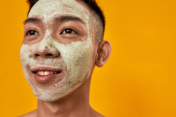 Close up portrait of young asian man with green mask applied on his face smiling away isolated over yellow background. Beauty, skincare routine concept