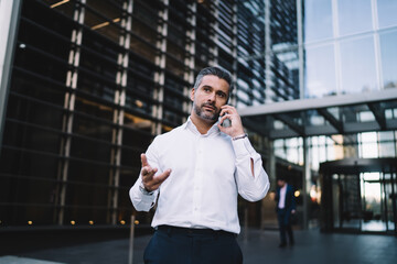 Formally dressed businessman using cellphone device for making international consultancy conversation at urbanity of financial district, mature banker in white shirt connecting to roaming for phoning