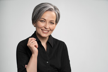 Charming gray haired woman wearing black shirt smiles at camera, isolated on white background. Mature Caucasian woman in studio raised her elbow bending arm. High quality photo.