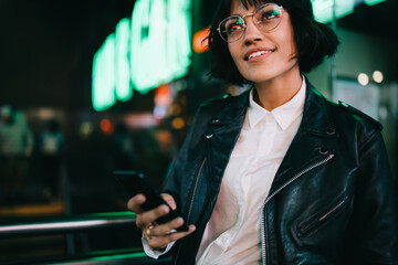Young brunette woman dressed in trendy leather jacket holding modern mobile technology and smiling,...