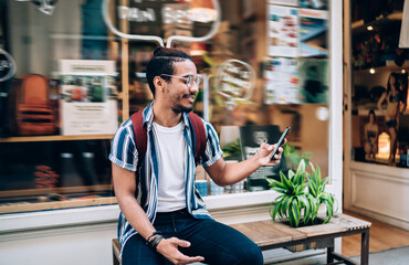 Smiling ethnic man browsing smartphone near shop