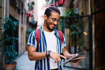 Smiling dark skinned male in eyewear for vision correction searching information strolling on...