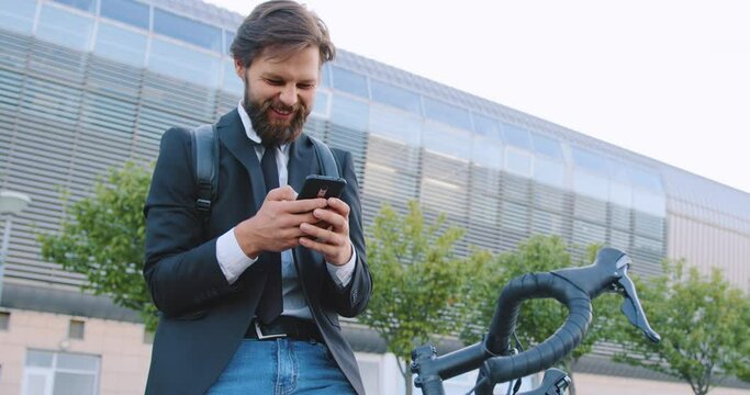 Front view of smiling handsome stylish bearded man which sitting on his bike and reviewing funny videos on his phone near beautiful modern city building