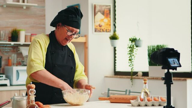 Mature experienced baker recording culinary recipe tutorial using modern camera in kitchen wearing bonete and apron. Influencer chef using internet technology communicating on social media.