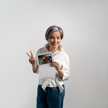 Woman with gray hair keeps colorchecker on an isolated background. Adult woman smiles and shows two fingers. High quality photo.