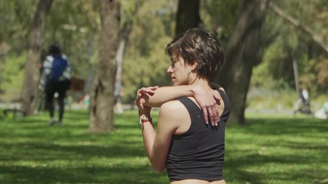 Medium shot of a pretty woman stretching her shoulders at a busy park. High quality 4k footage