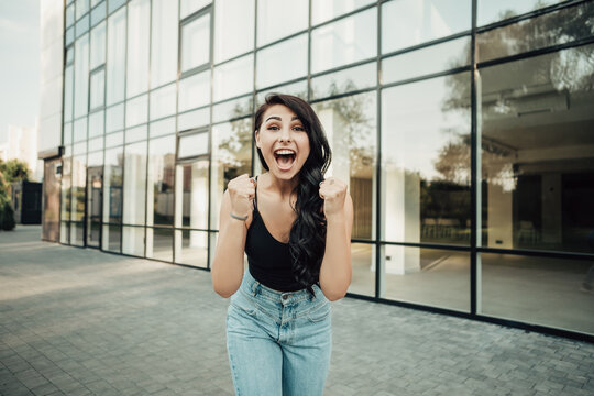 Portrait of excited happy young sensual woman screaming. Lady raises arms from joy, shouts loudly, dressed in singlet and jeans. Image with copy space. Celebration concept.