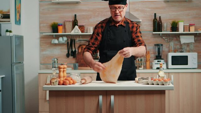 Chef cooking handmade pasta with healthy ingredients at home in modern kitchen. Happy elderly baker with bonete using wooden rolling pin, sprinkling, sieving flour on table, baking traditional cookies