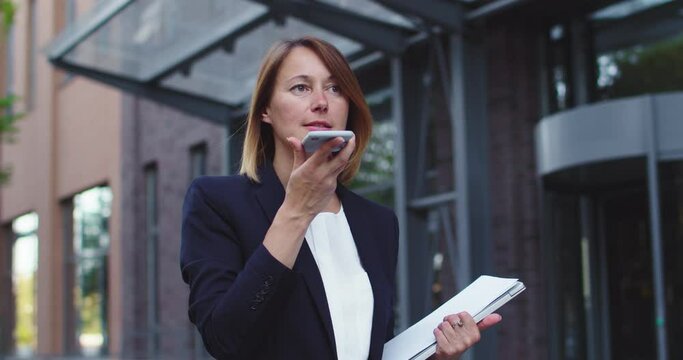 Attractive adult businesswoman in formal suit walking near modern office building. Caucasian female employee using smartphone to record voice messages to collegue. Technologies, Interner concept.