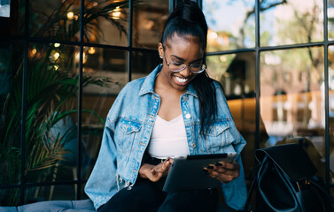 Cheerful dark skinned woman in eyewear for vision correction smiling checking salles in web store on digital tablet, positive african american hipster girl share media and multimedia content online