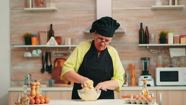 Woman wearing chef bonete while mixing cracked eggs with flour in kitchen while preparing food following traditional recipe. Retired elderly baker kneading in glass bowl ingredients for homemade cake