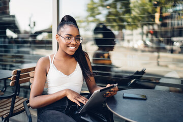 Portrait of cheerful dark skinned hipster girl in eyewear using digital tablet for searching information, happy african american woman blogger resting on free time on cafe terrace with touchpad