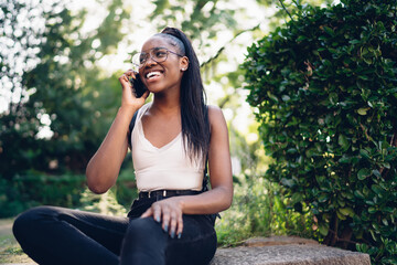 Cheerful dark skinned female sitting in city park relaxing on free time and laughing on smartphone conversation, positive 20s african american woman in spectacles and casual wear making roaming call
