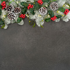 Traditional natural winter solstice snow covered spruce fir border with holly, mistletoe & pine cones on grey grunge background. Festive design for Christmas & New Year holiday season. Flat lay.
