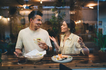 Multiethnic young couple having pleasant conversation while eating in cafe behind glass window