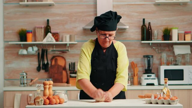 Grandmother preparing wheat homemade bread wearing chef bonete. Retired elderly baker with bonete mixing ingredients with wheat flour kneading dough for baking homemade cake and bread