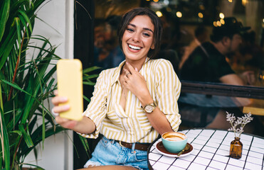 Joyful young ethnic woman taking selfie on smartphone during vacation in cafe