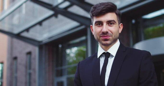 Portrait of attractive Caucasian male worker in formal suit near office building. Smart young businessman looking at camera and smiling outdoors. Occupation, profession, job, entreprenuership.