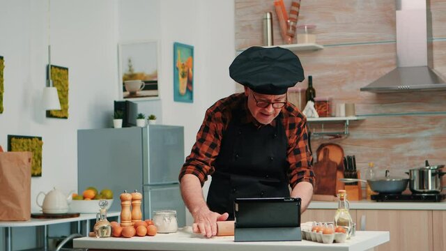 Retired man following culinary advice on tablet, learning cooking tutorial on social media, forming dough with wooden rolling pin. Grandfather with bonete and apron using laptop preparing home cakes