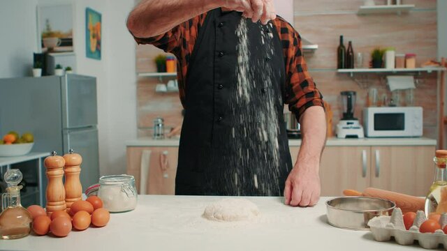 Bakery man sifting wheat flour, preparing bread dough. Retired elderly chef with bonete and uniform sprinkling, sieving spreading rew ingredients with hand baking homemade pizza and bread.
