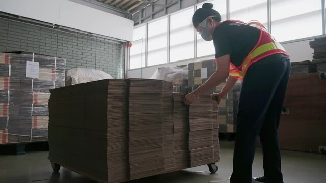 Asian woman factory worker doing arrangement cardboard paper to be stack for preparation process in warehouse room. Asia female wearing face mask while working in factory. New normal wear of people.