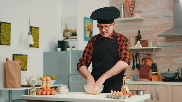 Baker kending dough in kitchen table wearing apron and bonete. Retired elderly chef with uniform sprinkling, sieving sifting raw ingredients by hand baking homemade pizza, bread.