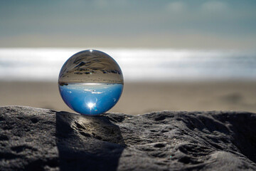 Photography Crystal Photo Ball showing upside down reflection of sky beach and waves balanced on rocks on sandy beach.