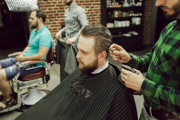 Haircut in the barbershop. Side view of young bearded man getting haircut while sitting in chair at barbershop. Hairdresser moisturizes hair with spray