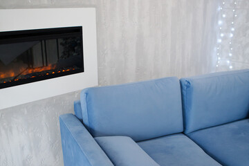 Blue sofa near modern closed fireplace with burning fire on gray wall with light bulbs garlands background. New Year's interior. copy space.