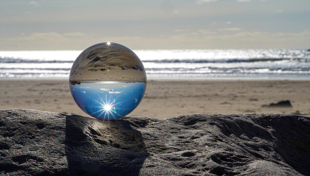 Photography Crystal Photo Ball showing upside down reflection of sky beach and waves balanced on rocks on sandy beach.