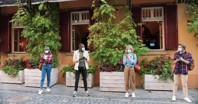 Mixed-races young people standing in line with smartphones in hands at social distance. Multi ethnic men and women in medical masks using gadgets and waiting in queue at street. Pandemic measures.