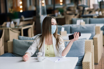 Serious woman with protective face mask looking at smart phone checking news on a cafe terrace