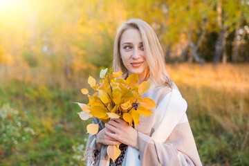 Young beautiful woman in long coat with yellow leaves basking with a blanket in autumn forest.