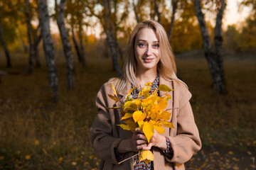 Young beautiful woman in long coat with yellow leaves posing in autumn forest.